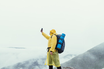 A male tourist in a yellow raincoat and with a backpack on his back stands on top of a mountain and catches a net on a smartphone.