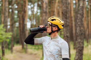 Handsome young man cyclist in helmet and goggles drinks water from a bottle on a background of pine...