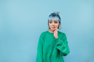 Cute shy hipster girl with colored hair speaks in secret and looks away on a blue background.
