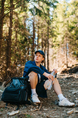 Fototapeta premium Handsome young man sitting in the woods with a backpack while hiking in the mountains with a bottle of water in his hands, looking away with a serious face.
