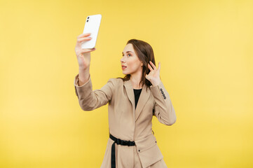 Naklejka premium Beautiful woman in jacket making selfie on yellow background, posing on smartphone camera, isolated.