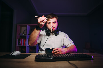 An adult man with a beard sits at the computer at night at work and eats Chinese food with...