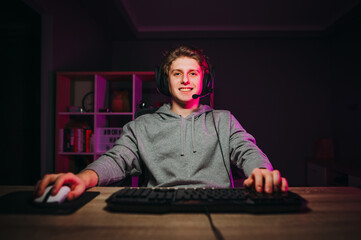 Positive young man in headset sitting at home at the computer and playing online games with a smile...