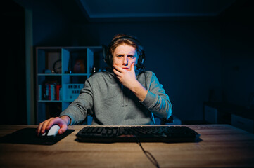 Concentrated young male gamer sleeping at night in a room at home in a headset and playing online games on the computer, looking with a serious face at the screen