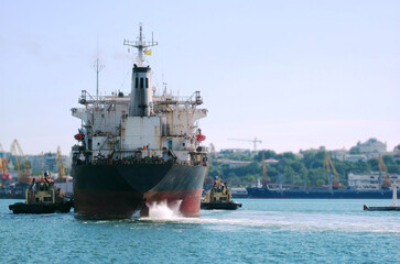 Obraz premium The ship enters the port. Two longboats are pushing the ship. Mushroom propellers foaming the water. A dry cargo ship's view of the back of the bay. Sunny Black Sea city in the background