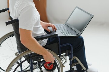 Obraz premium Portrait of smiling disabled male sitting in wheelchair and working on laptop from home. Young worker with special needs. Freelancer and people with disabilities concept.