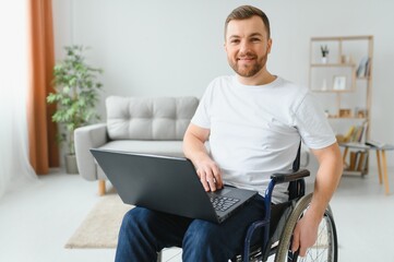 Portrait of smiling disabled male sitting in wheelchair and working on laptop from home. Young...