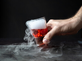 mano masculina sosteniendo Bebida roja con hielo seco sobre fondo negro y con base de piedra. reacción química con Humo saliendo