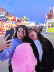 Two friends joking while taking selfie holding a cotton candy in a night fair