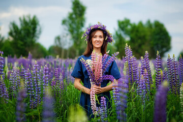 A young brunette in a flower wreath made of lupine with a bouquet of happiness smiles and looks at the camera in a flower meadow, inhales the fragrance of flowers.