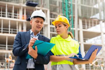Man engineer standing on a construction site with a young woman worker monitors the construction plan on a laptop, taking ..important notes