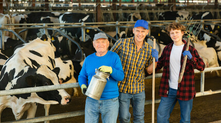 Three milk farm workers of different ages standing in cowhouse.