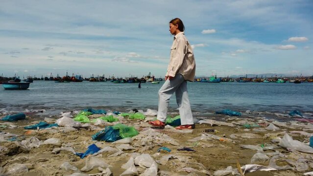 View of Caucasian girl walking on plastic bags shore. Fishing boats in background. Vietnam
