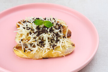 Pisang panggang, Grilled banana topped with chocolate sprinkles, grated cheese, and sweet white milk. Served on plate with mint leaves on grey background.
