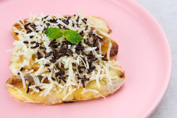 Pisang panggang, Grilled banana topped with chocolate sprinkles, grated cheese, and sweet white milk. Served on plate with mint leaves on grey background.
