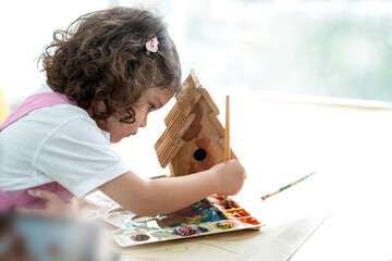 Cute little girl mixes colors to paint a wooden birdhouse in her room, education and learning...