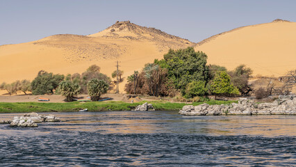 Boulders and green vegetation are visible on the banks of the Nile. Ripples on the blue water. Overturned boats lie on the sand. High dunes against the blue sky. Egypt