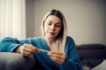 A sick woman looking at thermometer while sitting on sofa at her home.