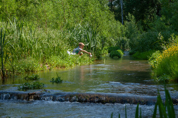 The boy sits on the bank of the river and sends paper boats to sail.