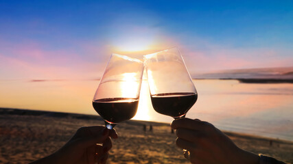 Close up of couple hands with red of glass wine in a holiday with wine glasses of couple in a beautiful sunset on beach  background