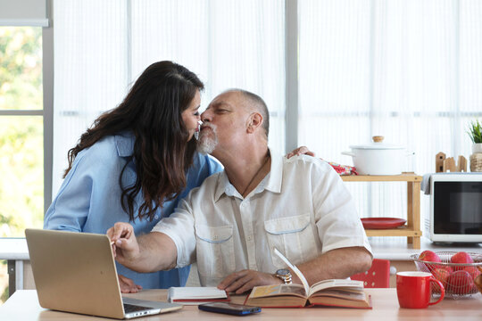 Romantic middle age couple having a sweet time together, woman morning kiss her husband while working with laptop in kitchen at home