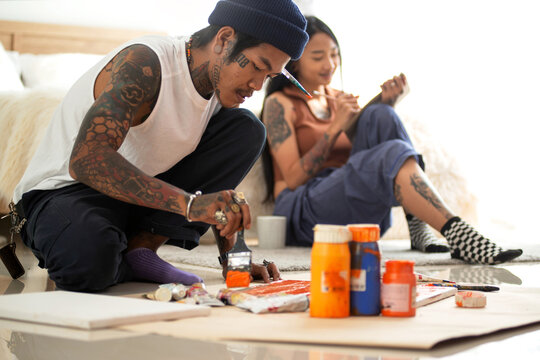 Man with tattoos on his body painted on a frame on the floor, girlfriend with tattoos reads a book in the background