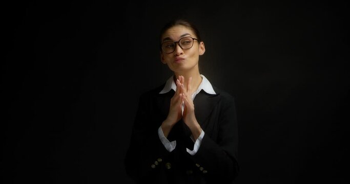 Cunning business woman in glasses and office clothes is standing on a isolated black background. Has evil plan in mind, steepled fingers, she came up with an idea