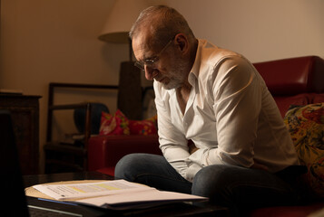 Adult man in white shirt siting on sofa at home and working with laptop in front