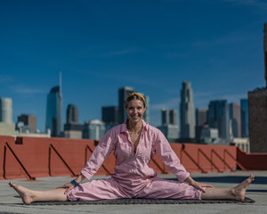 Fototapeta na wymiar Young blonde woman in pink coveralls and barefoot practices yoga on a downtown Los Angeles Roof top with the LA skyline behind her