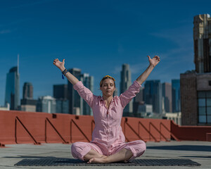 Fototapeta na wymiar Young blonde woman in pink coveralls and barefoot practices yoga on a downtown Los Angeles Roof top with the LA skyline behind her