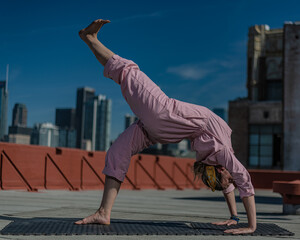 Young blonde woman in pink coveralls and barefoot practices yoga on a downtown Los Angeles Roof top with the LA skyline behind her