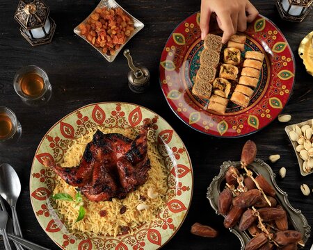 Female Hand Pick Turkish Baklava on Plate, Table Setting for Ramazan Greeting with Dates Fruit, Arabian Rice, ROasted CHicken, Nuts, and Tea.