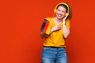 Laughing, smiling Girl in yellow basic suit and headphones holds small German flag. Study abroad...