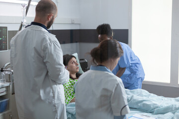 Patient admitted into the hospital recovering on bed recieving professional health care from experienced nurse while doctors examine symptoms. Team of medics consulting woman illness during rounds.