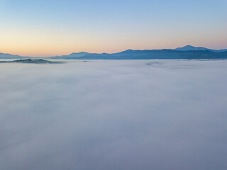 Naklejka premium Flight over fog in Ukrainian Carpathians in summer. Mountains on the horizon. A thick layer of fog covers the mountains with a continuous carpet. Aerial drone view.
