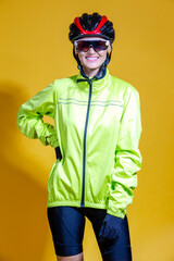 Female Road Cyclist in Professional Warm Outfit Standing In Green Autumn Jacket and Goggles With Reflections Against Yellow Background.