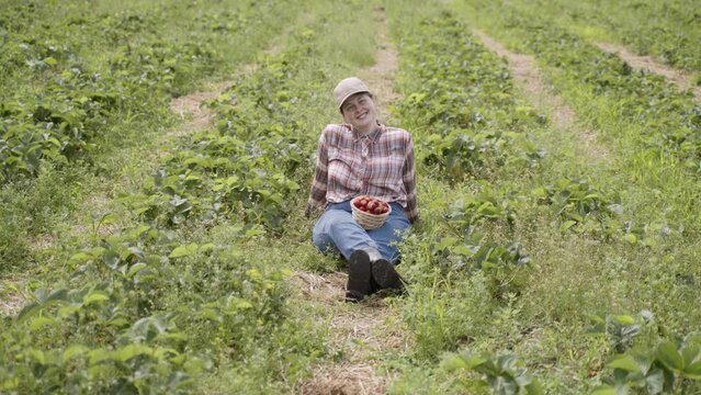 Happy farmer in plaid shirt and cap sits on path in strawberry field. On the feet of young woman stands basket of ripe red strawberries. The girl smiles and looks at the camera.