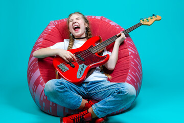 Caucasian Teenager Guitar Player With Red Shiny Bass Guitar Posing In Casual White Shirt With Smiling Facial Expression While Sitting on Cushion Chair On Turquoise Background.