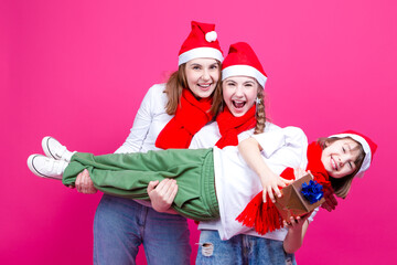 Fototapeta premium Group of Three Positive Caucasian Girls With Santa Hats Having Fun While Lifting Teenage Girl Holding Hands Closed Against Trendy Pink Background.