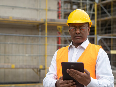 Indian engineer wearing safety vest and helmet looking down at his digital tablet computer