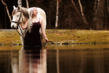 A young woman with white hair stands in the water with a white horse and turns her head