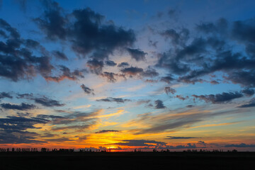 A bright, colorful sunset in the countryside. An orange-blue sky with clouds hangs over a field. A beautiful sundown.