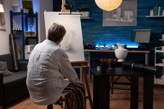 Senior woman feeling inspired looking at creative pencil drawing of vase on easel sitting down in professional art studio. Elderly artist in the evening finshing shading black and white sketch.