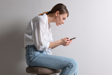 Woman with bad posture using smartphone while sitting on stool against light grey background - obrazy, fototapety, plakaty
