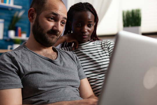 Young Adult Interracial Couple Looking At Computer Screen While Checking Social Network Feed. People Websurfing On Modern Laptop While Sitting Together On Sofa In Living Room At Home