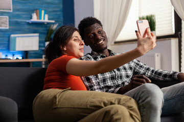 Joyful multiethnic couple posing for selfie sitting on sofa at home. Casual happy adult people bonding while taking selfies for fun using smartphone device in living room