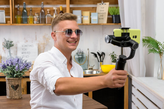 Young Handsome Guy Filming A Video Blog On A Mobile Device Using An Electronic Image Stabilizer In The Kitchen. Blond European Man Shoots A Video In A Good Mood