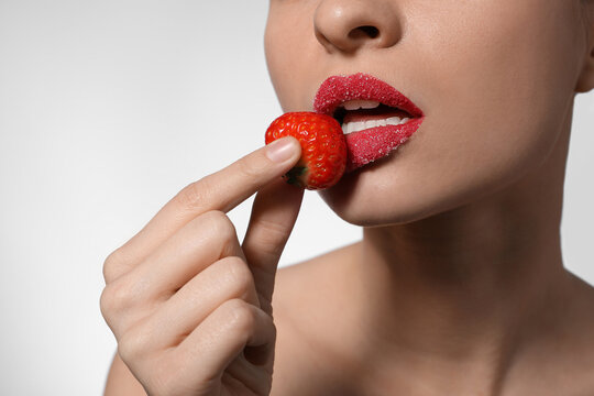 Young woman with lips covered in sugar holding strawberry on light background, closeup