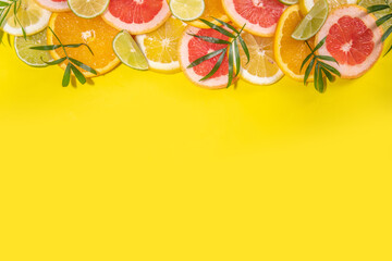 Colorful flatlay of citrus fruit slices and tropical palm leaves. Orange, grapefruit, lemon, lime bright high-colored slices on yellow background. Summer holiday background top view
