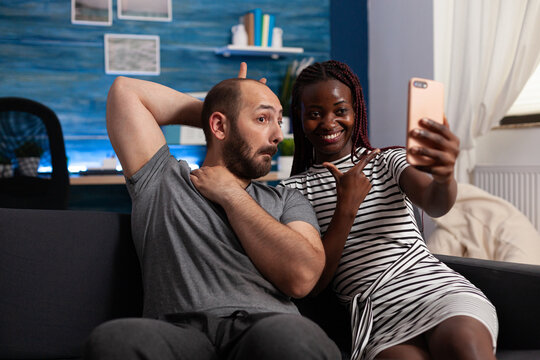 Boyfriend being goofy while joyful girlfriend taking selfie using smartphone. Cheerful african american wife and caucasian husband taking pictures together with phone while sitting on sofa in living